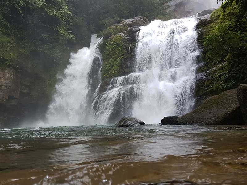 Um homem de pé num rio junto a uma cascata.