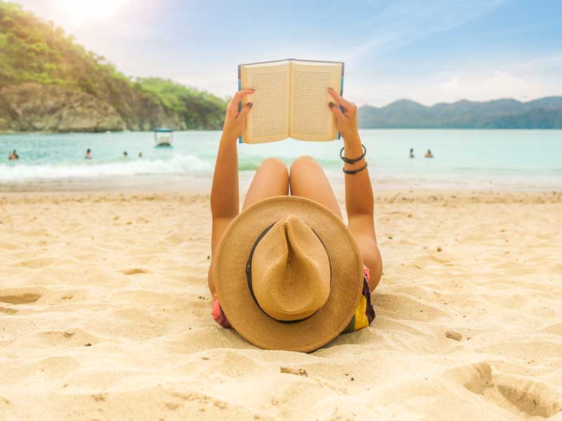 Uma mulher deitada na praia a ler um livro.