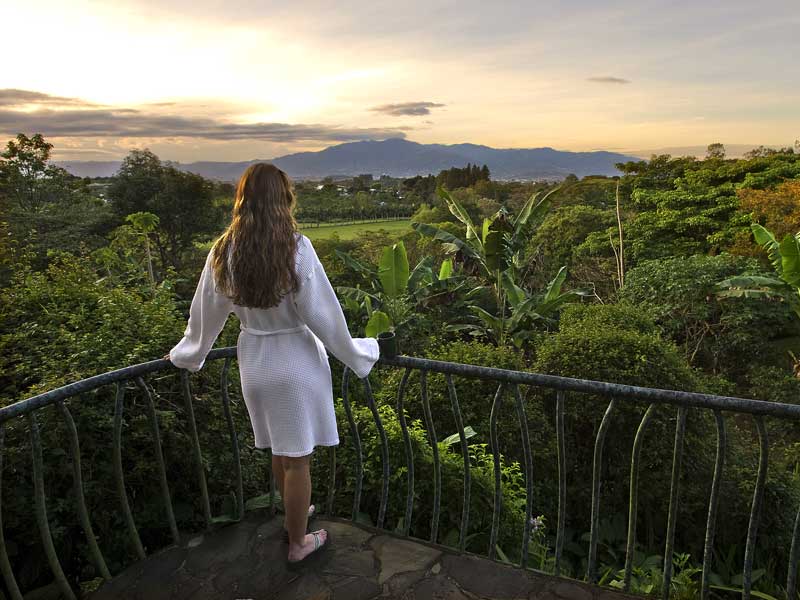 Uma mulher de pé numa varanda com vista para um vale verdejante.