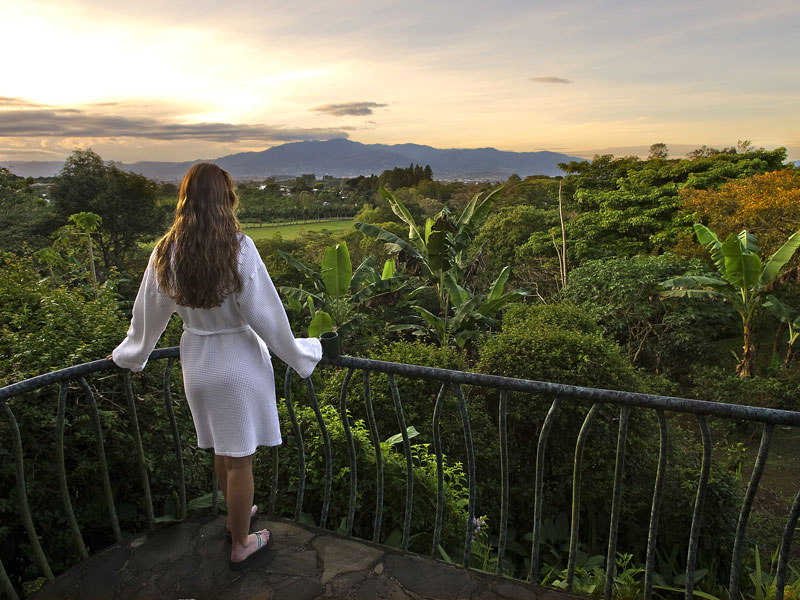Uma mulher de pé numa varanda com vista para um vale verdejante.