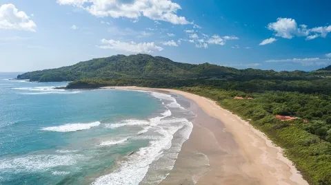 Vista aérea de uma praia de areia ao longo da costa do Pacífico d Costa Rica, rodeada por águas turquesa e colinas verdejantes.