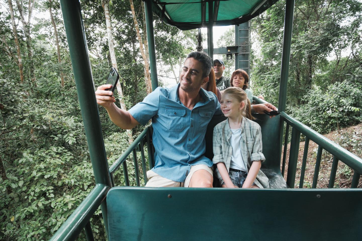 Uma família a tirar uma selfie numa gôndola ao ar livre, acima da copa da floresta tropical.