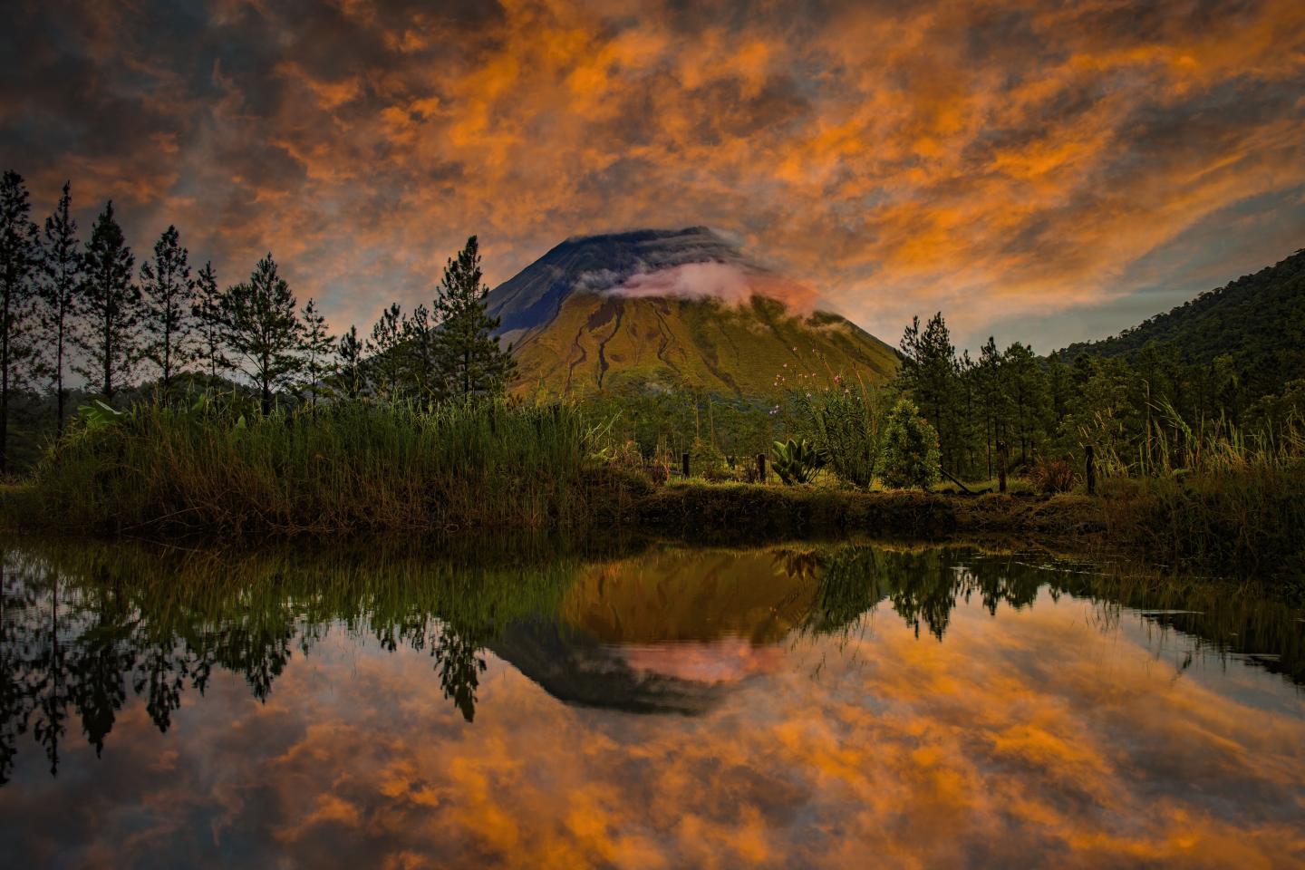 Um vulcão ergue-se sob as nuvens alaranjadas do pôr-do-sol, refletindo-se numa lagoa tranquila.