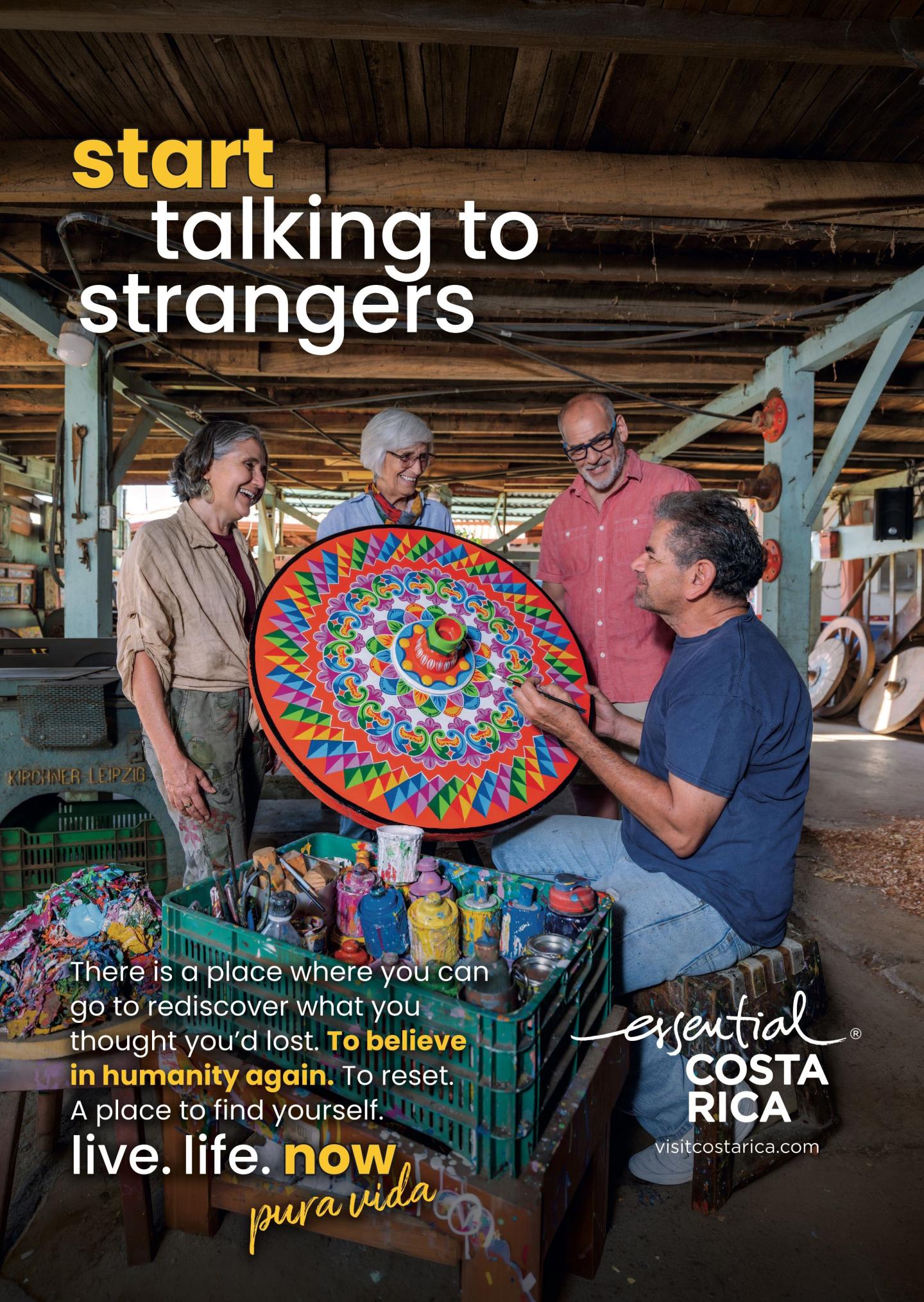 Anúncio que mostra quatro pessoas numa oficina a conversar com um artesão que segura uma peça artesanal circular, grande e pintada com cores vivas. Título: «Comece a falar com estranhos». Marca: «esencial Costa Rica, visitcostarica.com.
