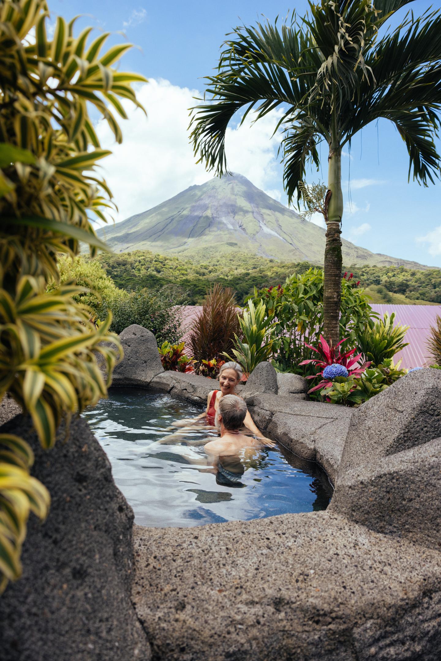 Duas pessoas relaxam numa piscina termal de pedra, rodeada por plantas tropicais, com o Vulcão Arenal ao fundo.