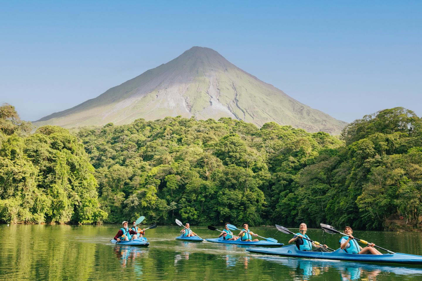Passeio de caiaque em grupo num lago tranquilo rodeado por floresta tropical, com o Vulcão Arenal a erguer-se ao fundo.