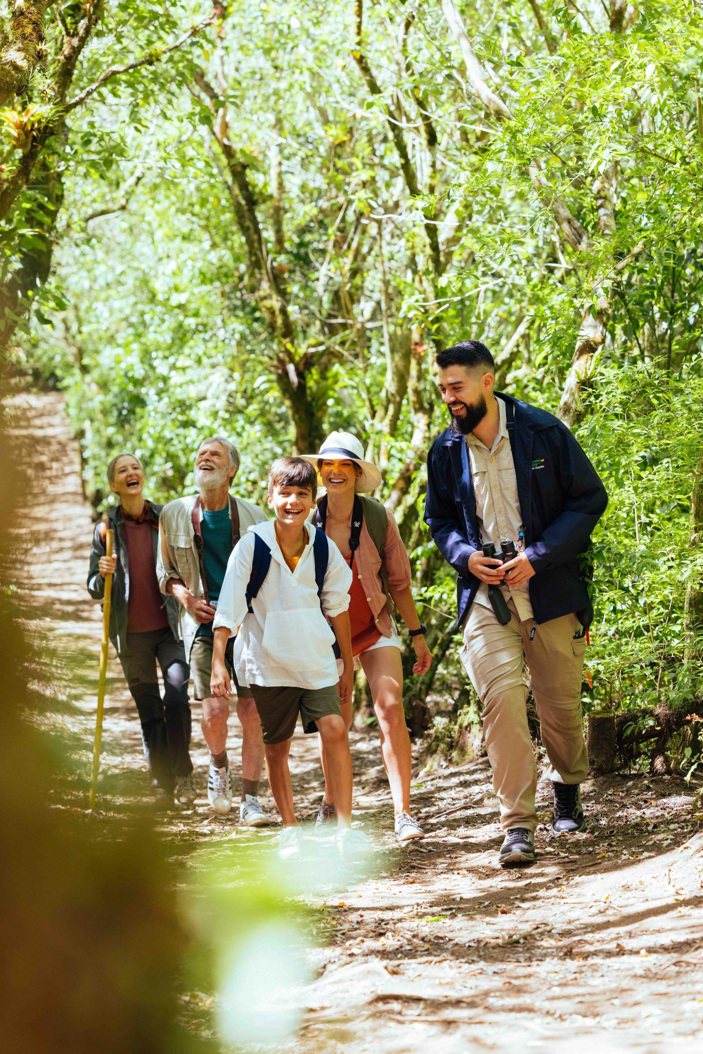 Um pequeno grupo de viajantes caminha por um trilho florestal à sombra, acompanhado por um guia.